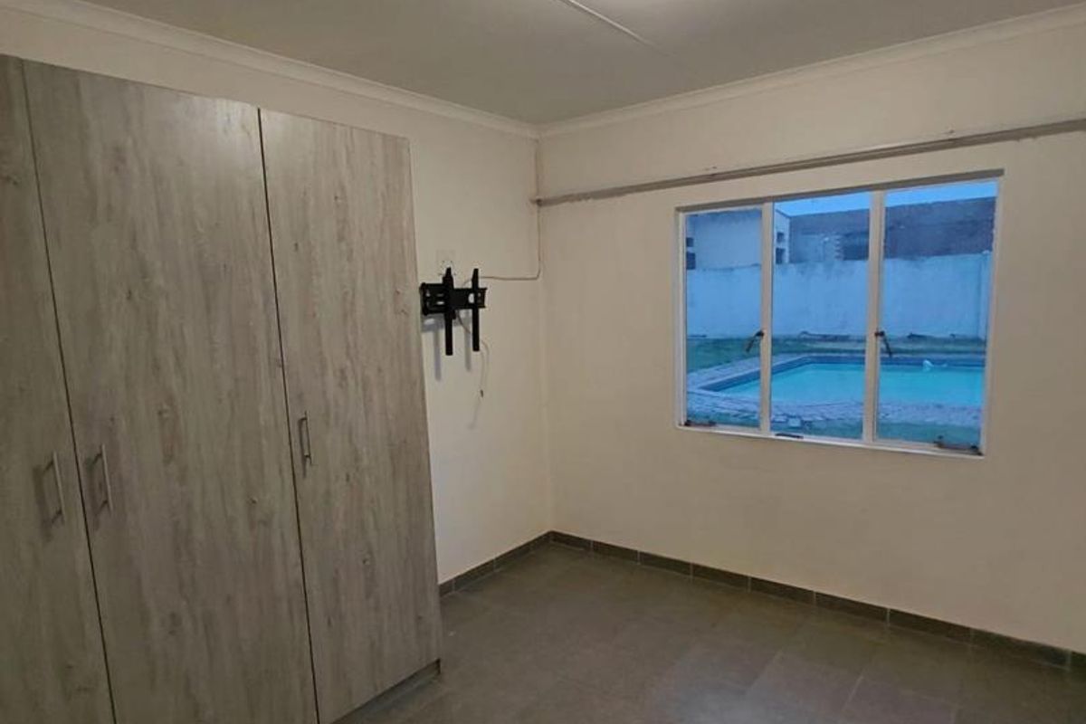 A clean, empty bedroom featuring built-in cupboards, tiled flooring, and a window view overlooking the swimming pool area.