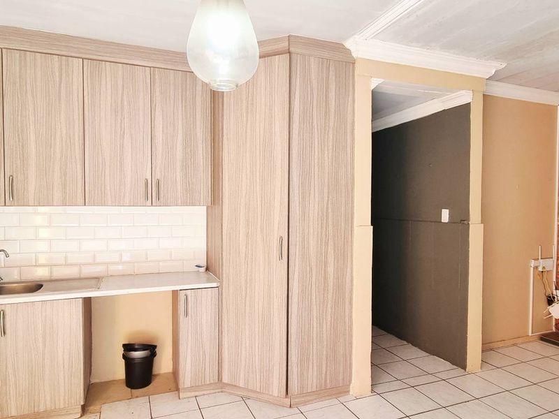 A compact kitchen featuring light wood-grain cabinetry, a subway tile backsplash, and a double sink, adjacent to an exposed brick wall and tiled flooring.