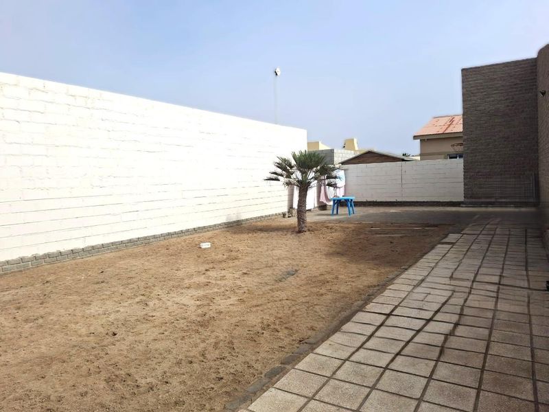 A rear exterior space featuring a paved walkway, a large sandy garden area, and perimeter boundary walls.