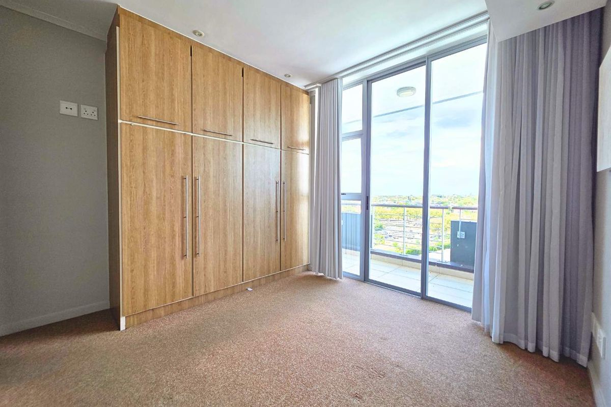 A bright, empty bedroom featuring floor-to-ceiling built-in wooden cupboards, carpeted flooring, and a large sliding glass door leading to a balcony.