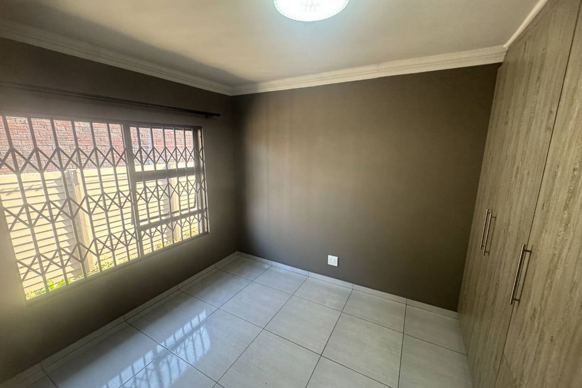 A clean, empty bedroom featuring neutral wall paint, tiled flooring, built-in wooden cupboards, and a window fitted with security bars.
