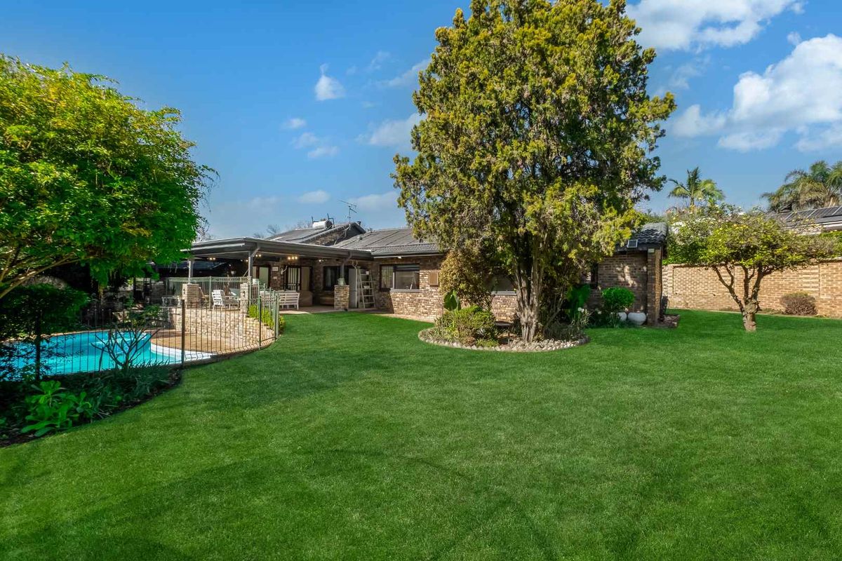 Spacious rear garden featuring a well-maintained lawn, mature trees, a swimming pool with safety fencing, and a covered patio area.
