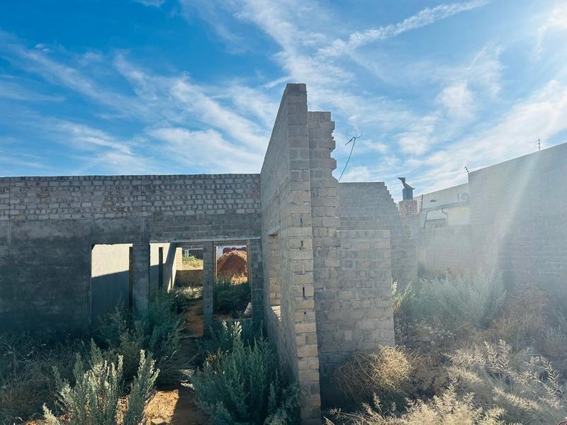 An unfinished residential structure featuring exposed brick masonry walls in a state of early construction, surrounded by overgrown vegetation.