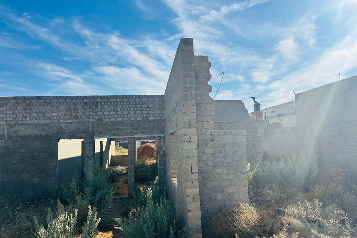 An unfinished residential structure featuring exposed brick masonry walls in a state of early construction, surrounded by overgrown vegetation.