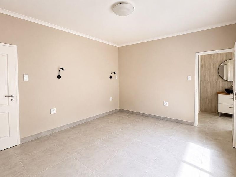 A bright, neutral-toned bedroom featuring tiled flooring, wall-mounted lighting, and direct access to an en-suite bathroom.