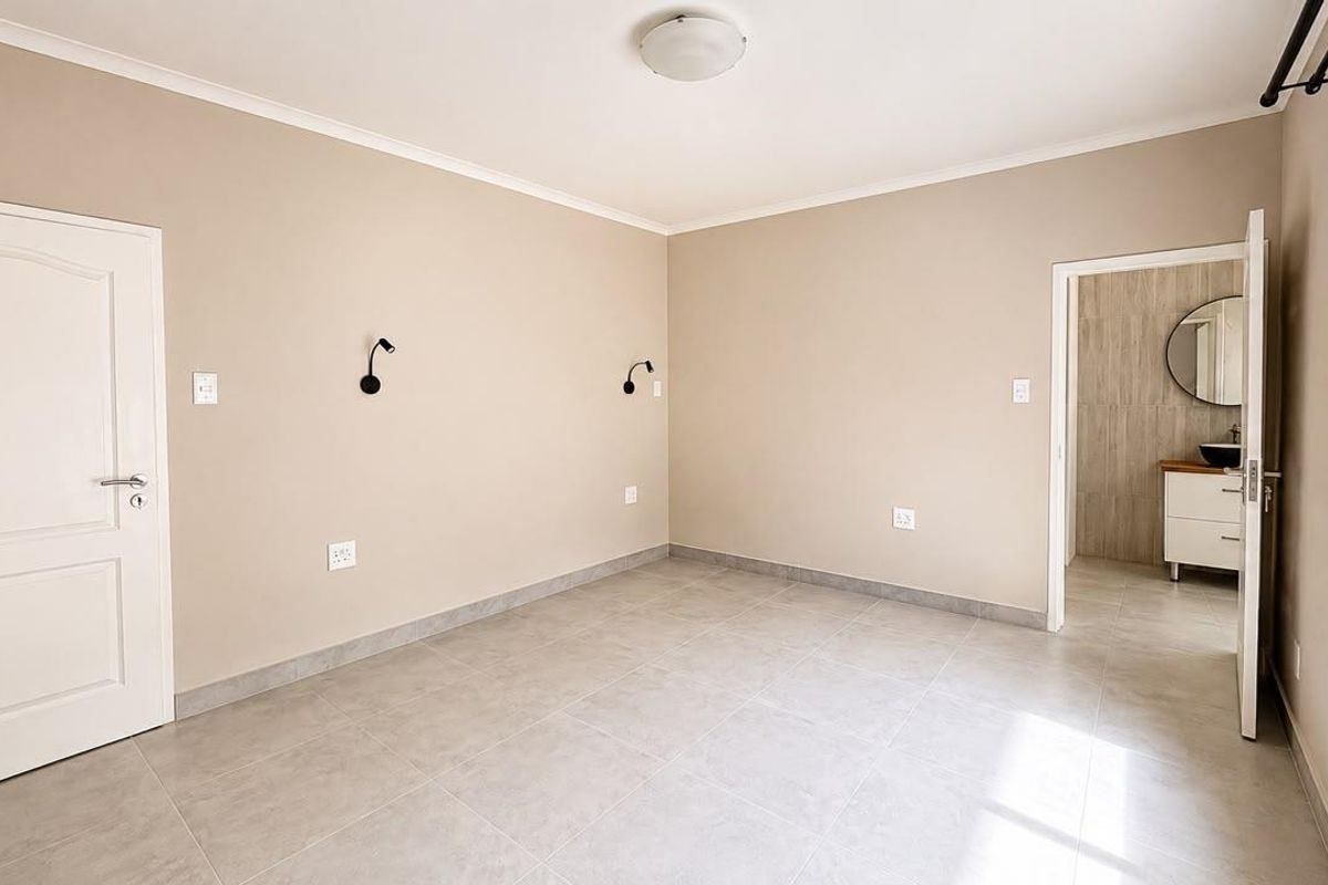 A bright, neutral-toned bedroom featuring tiled flooring, wall-mounted lighting, and direct access to an en-suite bathroom.