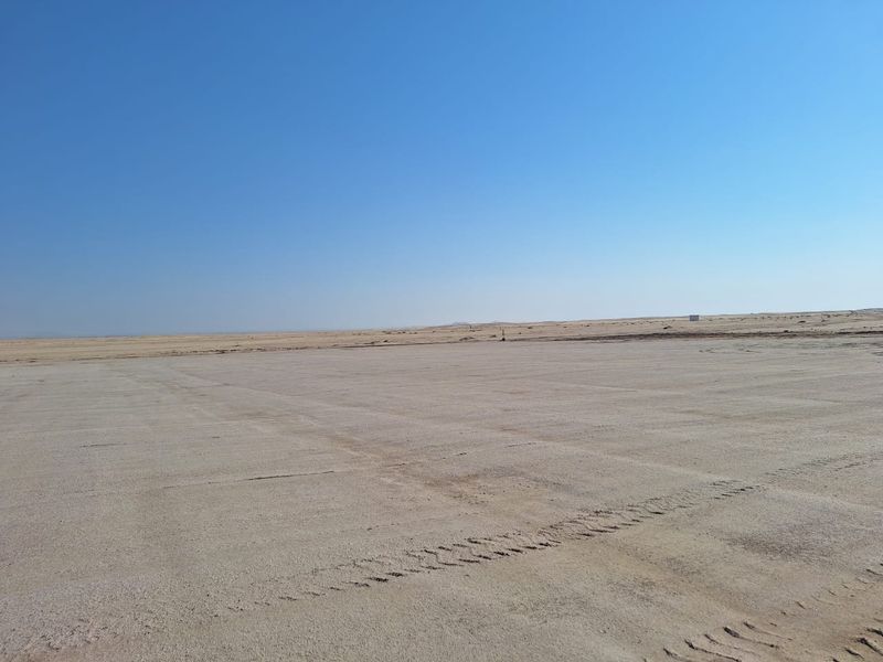 A wide, open expanse of undeveloped industrial land with a flat, sandy surface under a clear blue sky.
