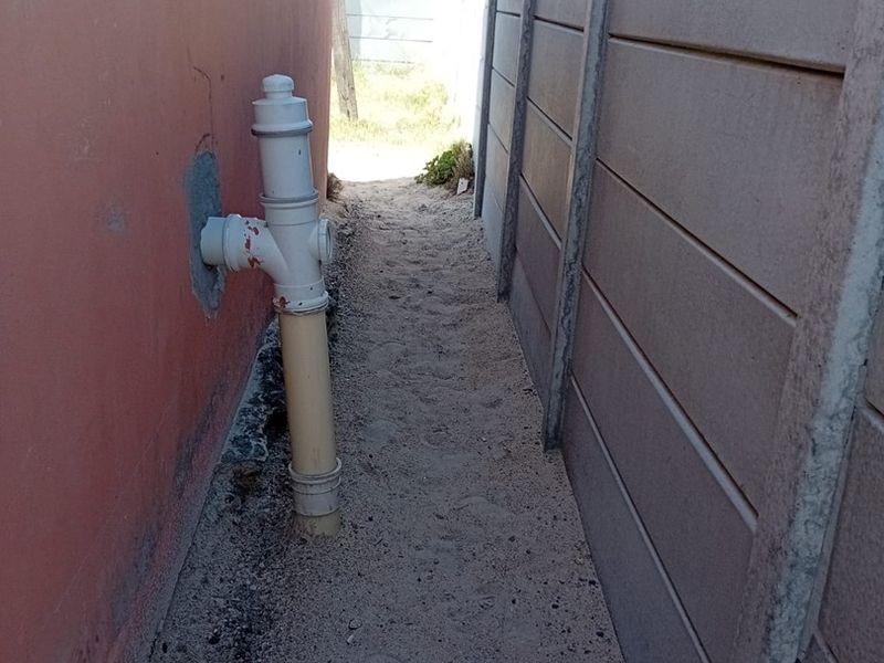 A narrow side passage between the house exterior wall and a precast concrete boundary wall, featuring exposed plumbing and an unpaved sandy surface.