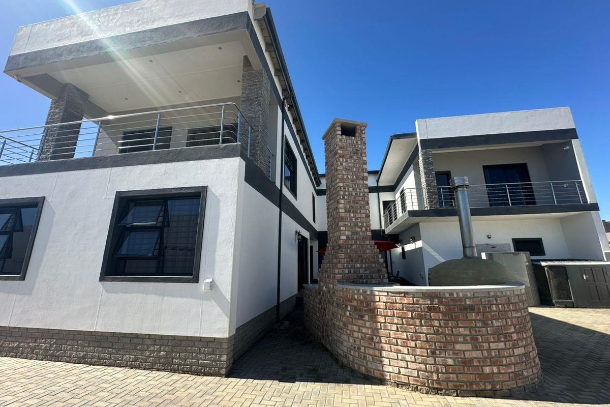 Rear exterior view of a modern two-story house featuring a brick outdoor fireplace, pizza oven, paved courtyard, and upper-level balconies with stainless steel railings.