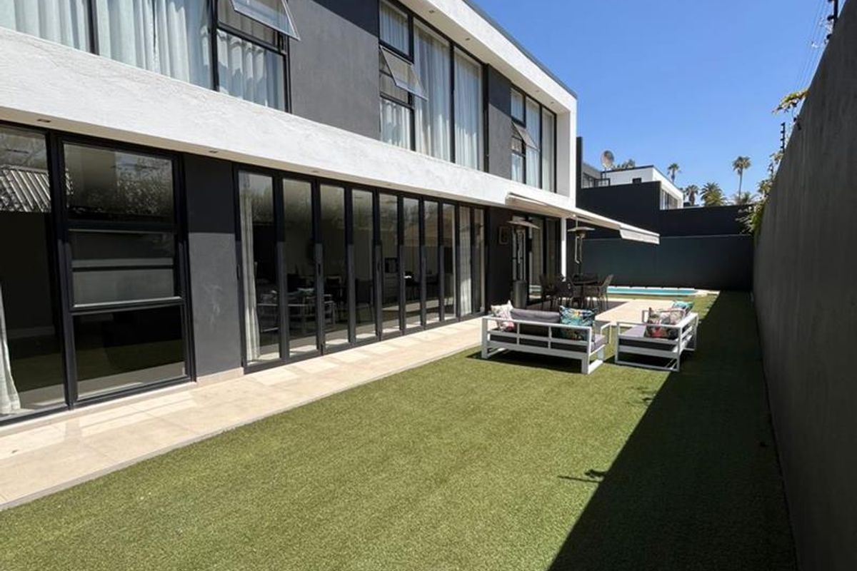 Modern rear exterior featuring a manicured artificial lawn, large bi-fold doors leading to the interior, a retractable awning, and a private swimming pool.