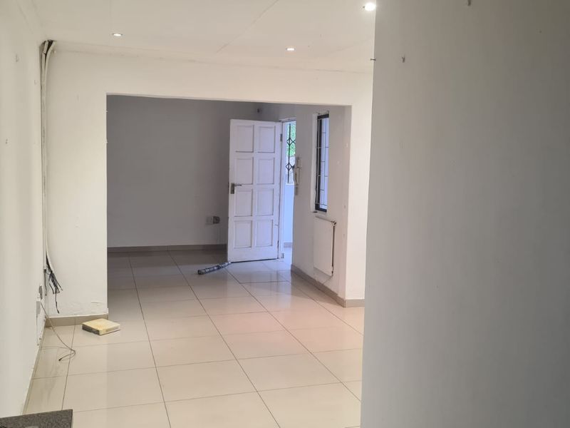 An empty, open-plan living area featuring neutral tiled flooring, recessed lighting, and a radiator, presenting a blank canvas for renovation.