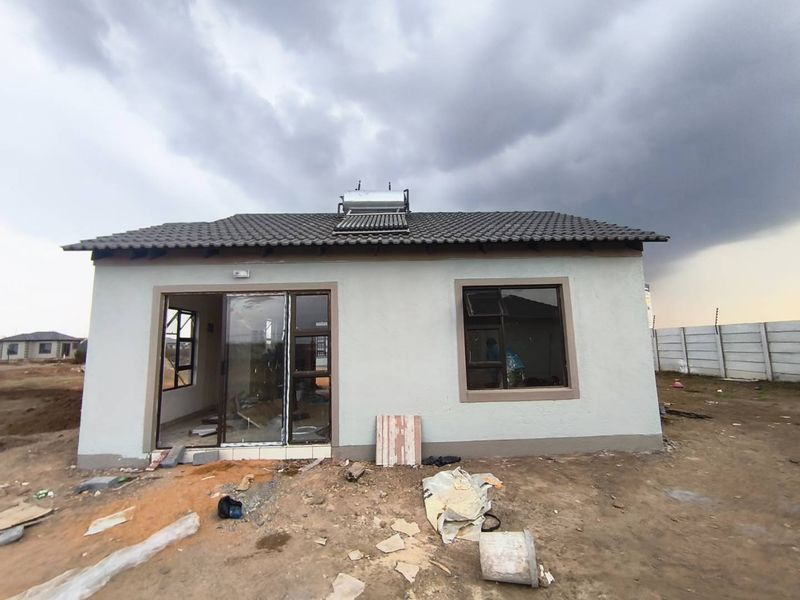 Front view of a newly constructed house featuring a tiled roof, solar water heater, and aluminium window frames, set on an unfinished plot.