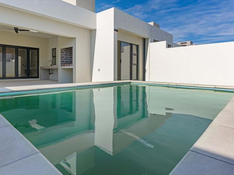 A modern outdoor entertainment area featuring a swimming pool, covered patio with a built-in braai, and sleek architectural lines.