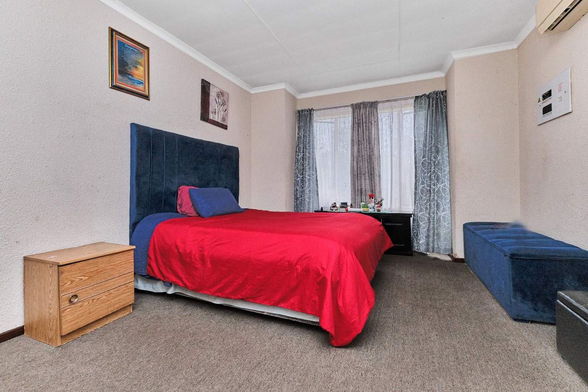 A standard bedroom featuring carpeted flooring, a wall-mounted air conditioning unit, and large windows allowing for natural light.