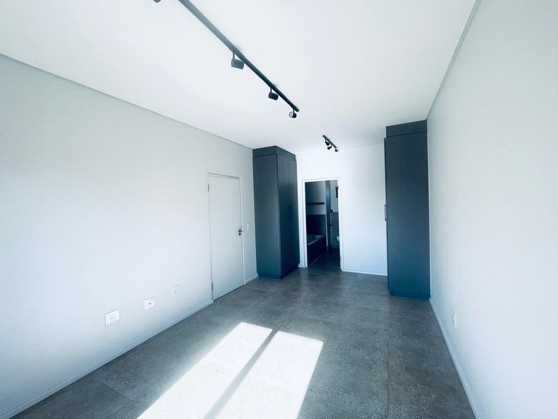 A modern, empty bedroom featuring sleek dark built-in cupboards, contemporary track lighting, and neutral tiled flooring, leading to an en-suite bathroom.