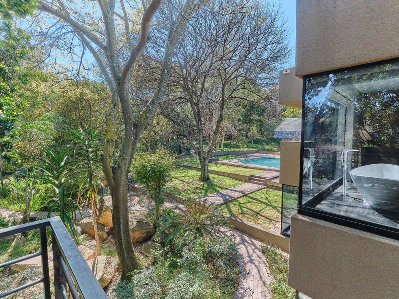 Elevated view overlooking a lush landscaped garden with mature trees, paved pathways, and a swimming pool, viewed from a modern balcony.