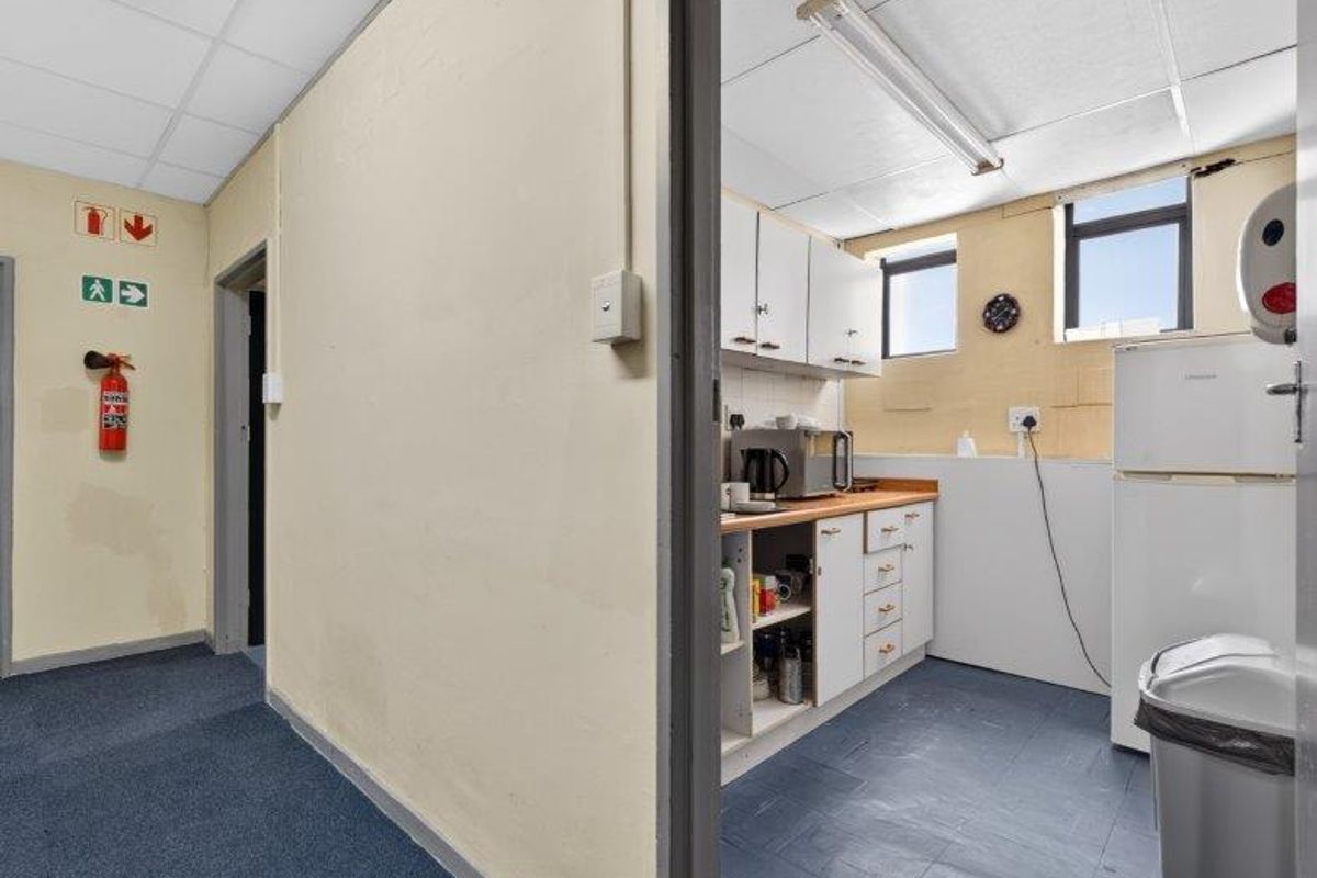 A functional office kitchenette featuring built-in cabinetry, laminate countertops, and a drop ceiling, adjacent to a carpeted hallway with safety signage.