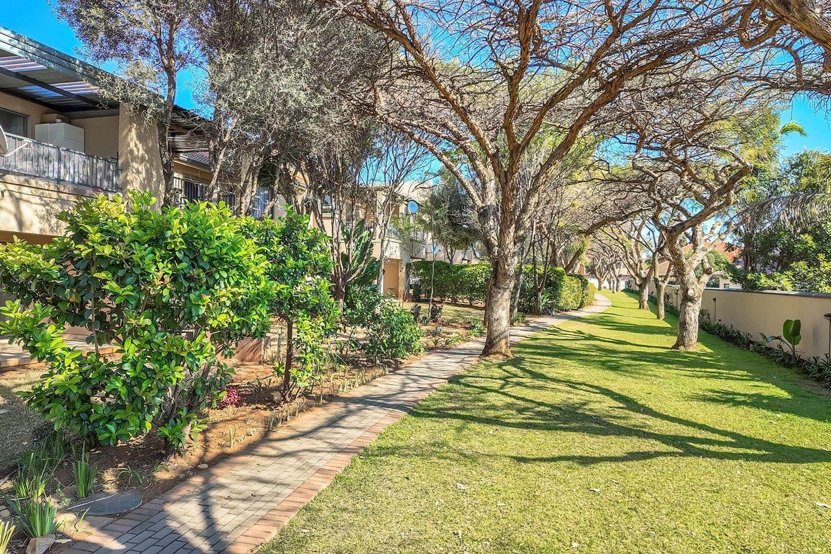 A well-maintained communal garden area featuring a paved walkway, mature trees, and manicured lawns adjacent to the apartment building's rear patio.