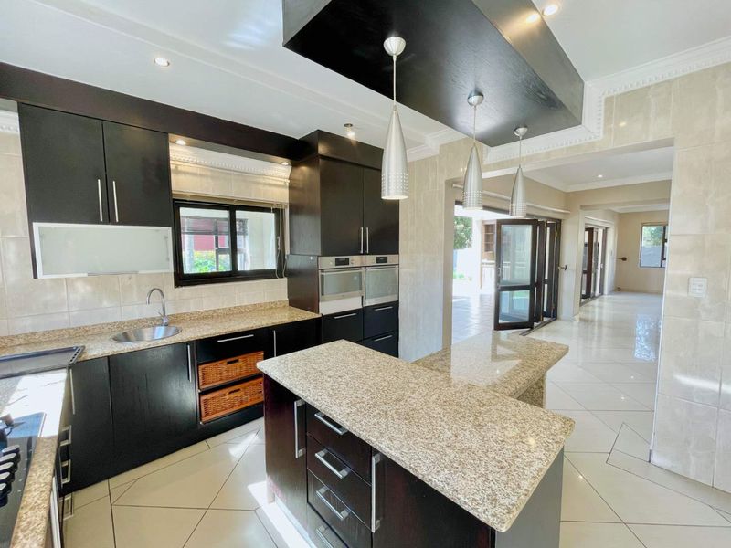 Modern kitchen featuring dark cabinetry, granite countertops, a central island, built-in double ovens, and elegant pendant lighting, flowing into an open-plan living area.