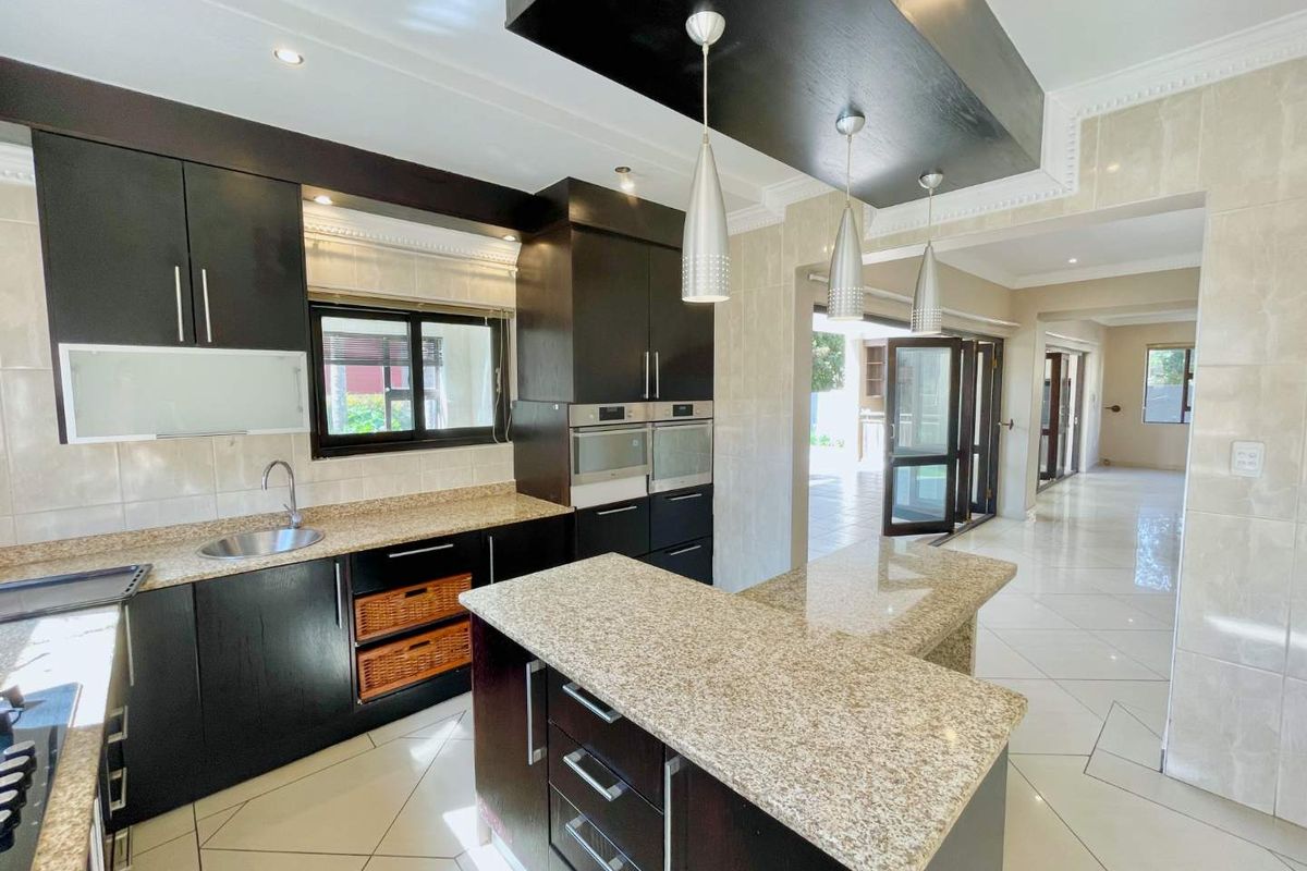 Modern kitchen featuring dark cabinetry, granite countertops, a central island, built-in double ovens, and elegant pendant lighting, flowing into an open-plan living area.