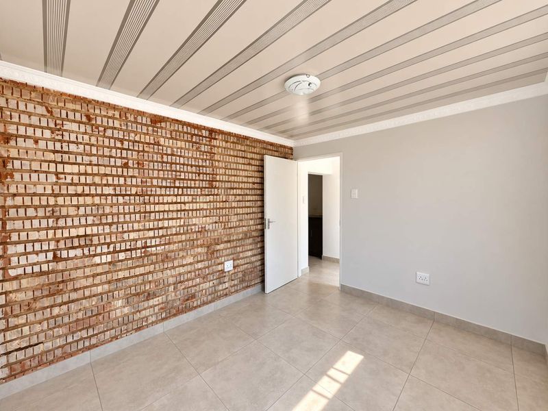 A bright, empty bedroom featuring a rustic exposed brick feature wall, neutral tiled flooring, and a unique striped ceiling design.