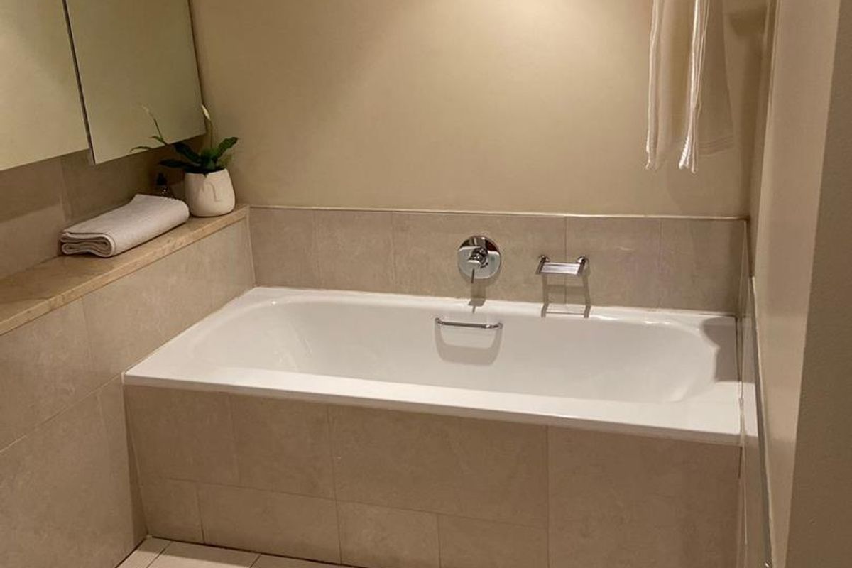 A clean, neutral-toned bathroom featuring a built-in bathtub with tiled surround, wall-mounted mixer, and integrated mirror cabinetry.