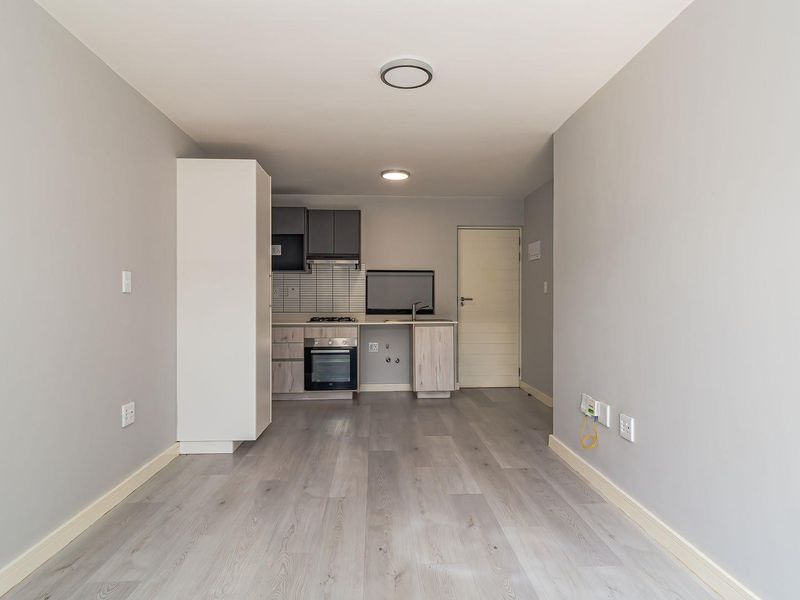 An open-plan living area featuring a modern kitchenette with built-in oven, hob, and cabinetry, finished with light-toned laminate flooring.