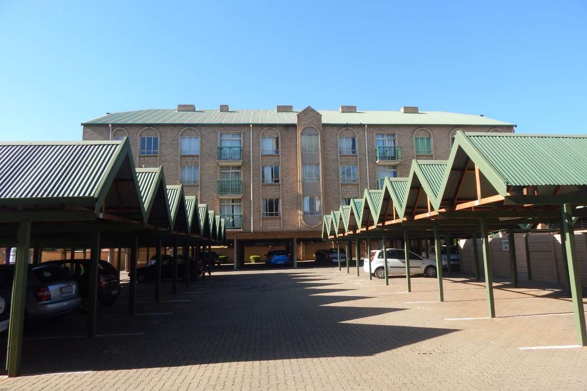 Exterior view of a multi-story apartment building featuring a brick facade, arched windows, and extensive covered parking bays with corrugated metal roofing.