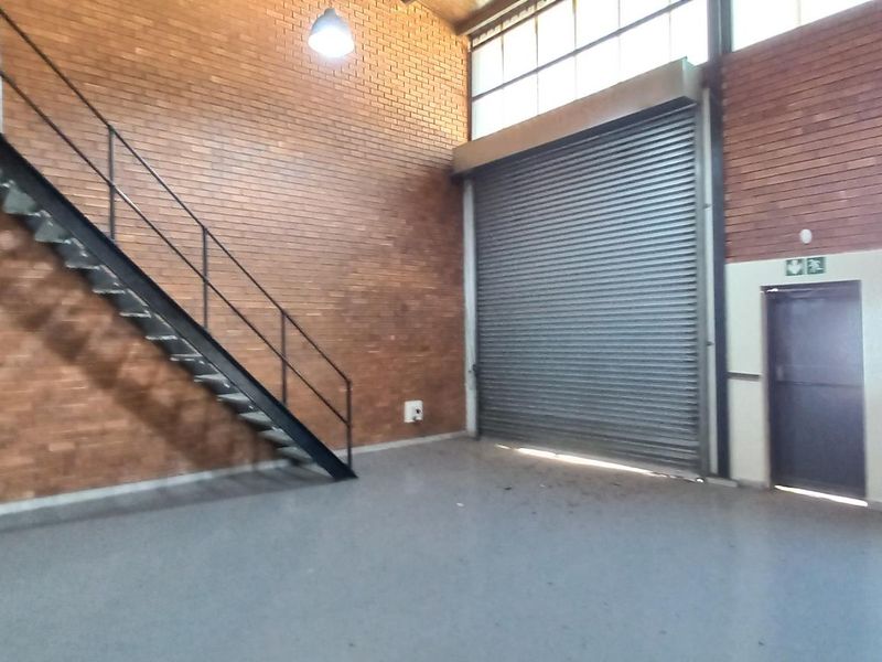 Spacious industrial warehouse unit featuring exposed brick walls, a high-clearance roller shutter door, and a metal staircase leading to an upper level.