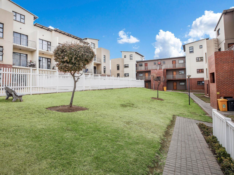 A well-maintained communal garden area within an apartment complex, featuring paved walkways, manicured lawns, and modern multi-story residential buildings with balconies.