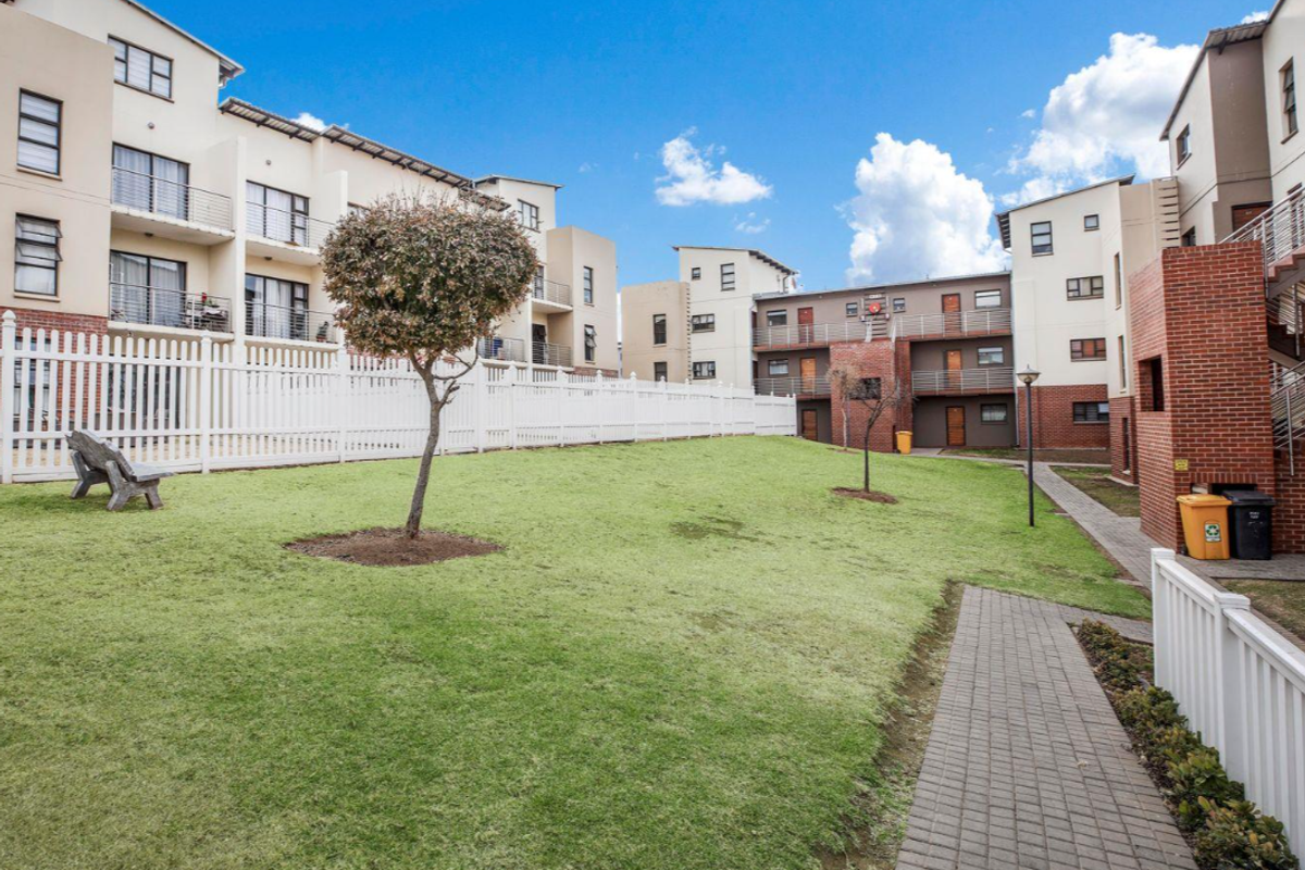 A well-maintained communal garden area within an apartment complex, featuring paved walkways, manicured lawns, and modern multi-story residential buildings with balconies.