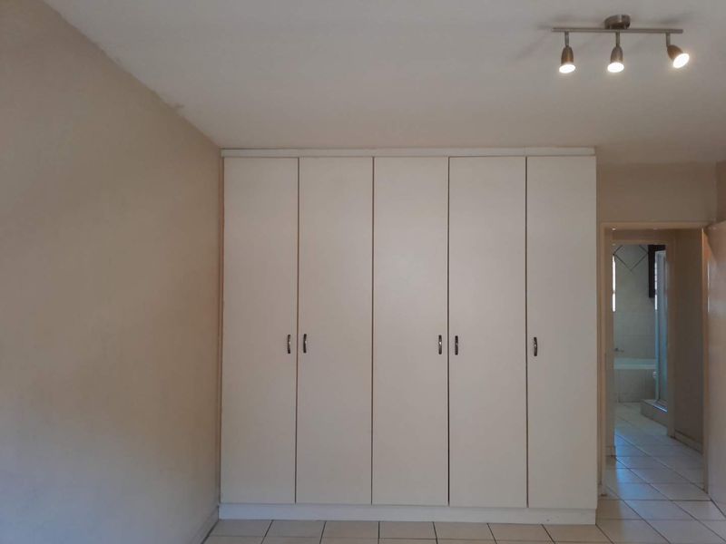 A clean, empty bedroom featuring neutral tiled flooring, built-in white cupboards, and modern track lighting, with an adjacent view into a tiled bathroom.