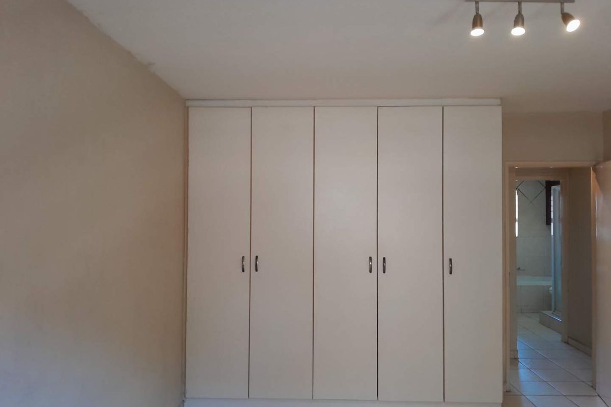 A clean, empty bedroom featuring neutral tiled flooring, built-in white cupboards, and modern track lighting, with an adjacent view into a tiled bathroom.