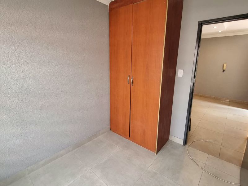 A clean, empty bedroom featuring neutral grey walls, light-toned tiled flooring, and a built-in wooden wardrobe.