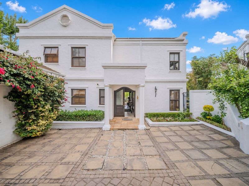 A well-maintained double-story home featuring a paved front courtyard, established garden beds, and a covered entrance with classic architectural detailing.