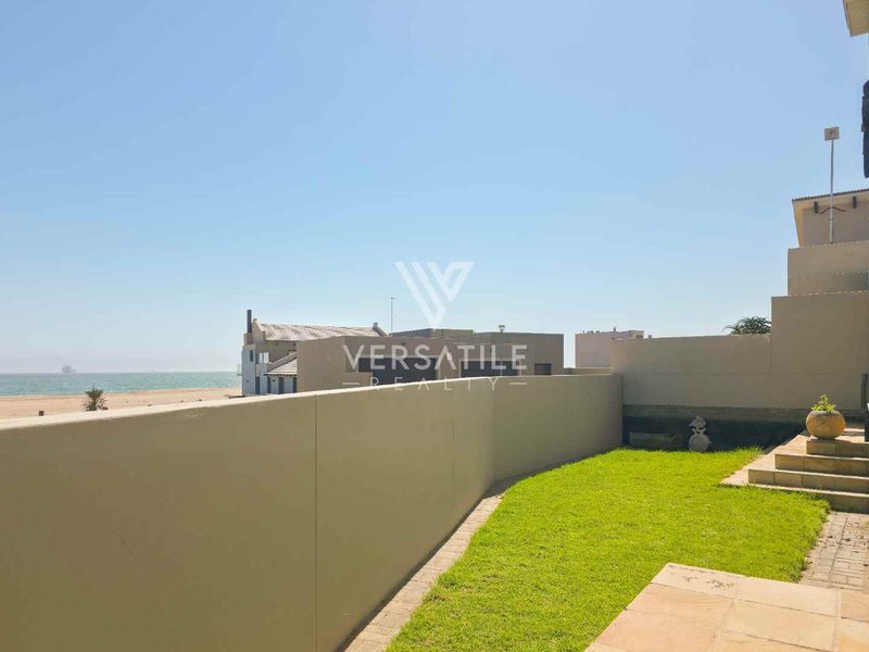 A rear exterior view of the townhouse featuring a private lawn, paved patio area, and boundary wall with direct access to the beach.