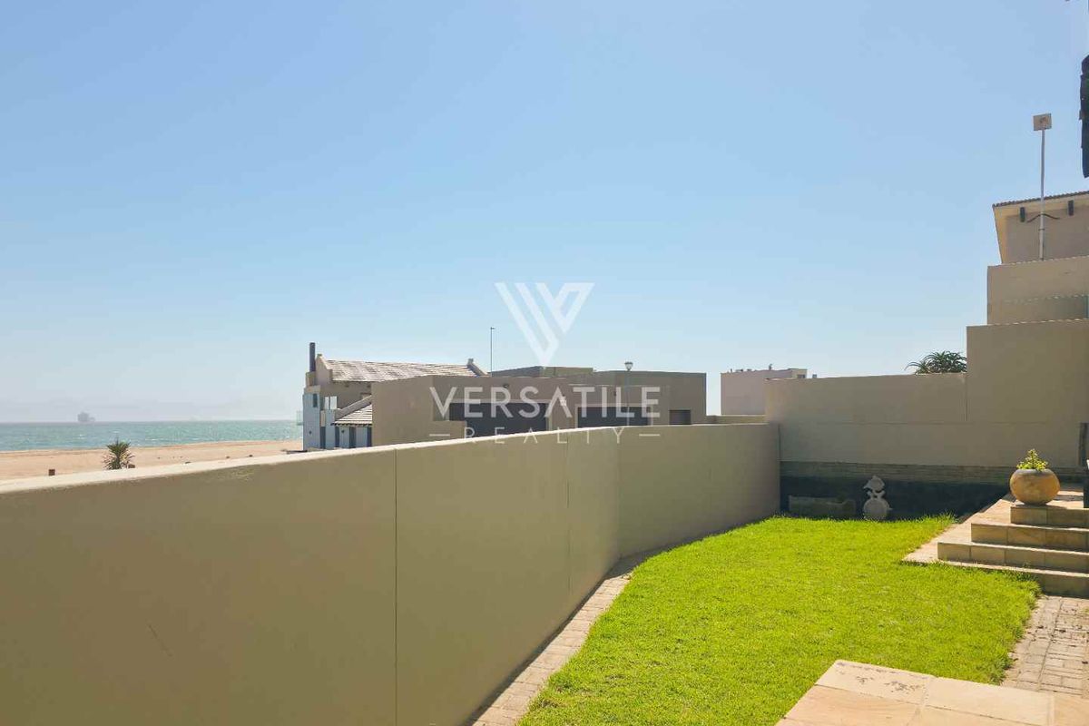 A rear exterior view of the townhouse featuring a private lawn, paved patio area, and boundary wall with direct access to the beach.
