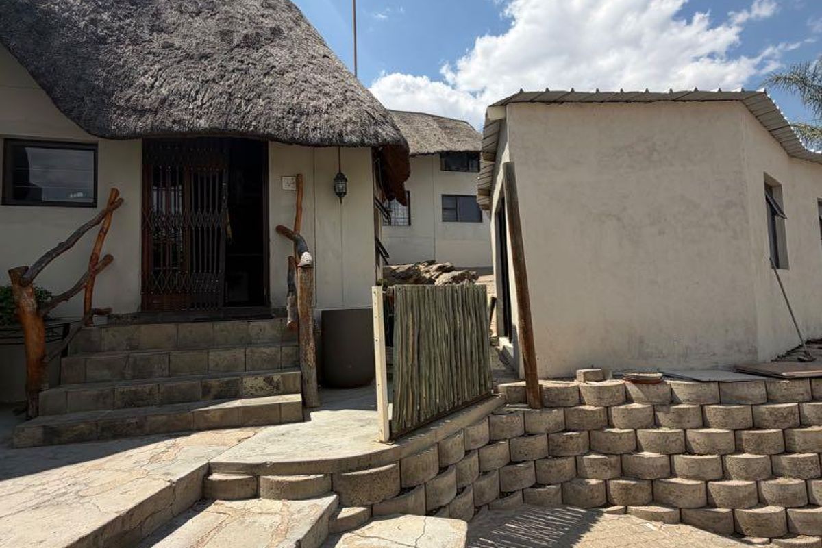 Rear exterior view featuring a thatched roof structure, stone retaining walls, and paved patio area with steps leading to the entrance.