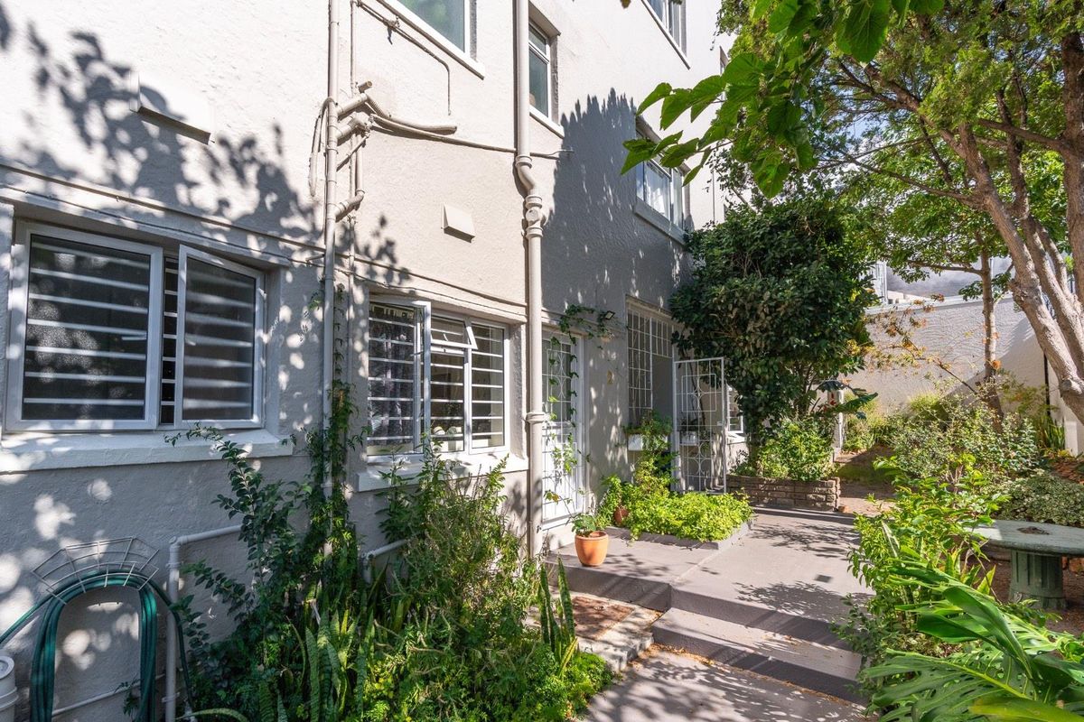 A view of the apartment's rear exterior, featuring a paved walkway, established garden landscaping, and secure window grilles.