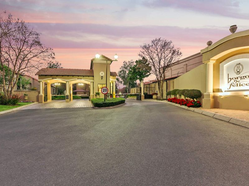 The secure entrance gatehouse of Borgo de Felice apartment complex, featuring paved driveway access, perimeter walls, and well-maintained landscaping.