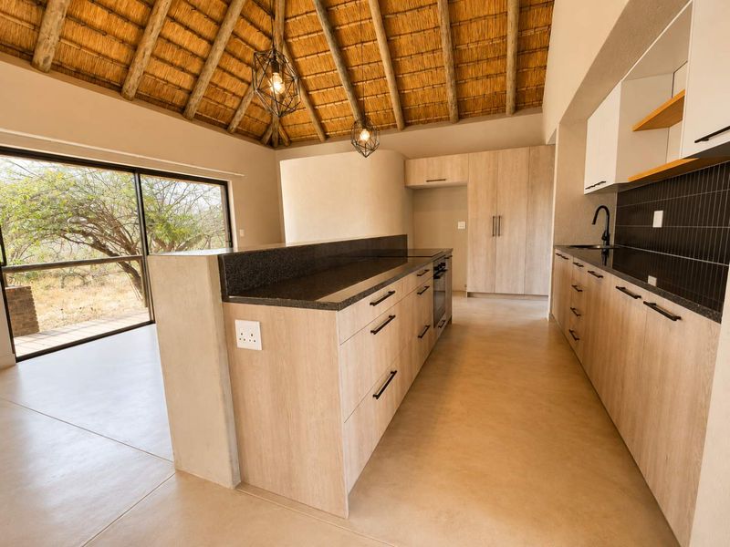 Modern kitchen featuring light wood cabinetry, dark stone countertops, and a high thatched ceiling, opening onto a scenic outdoor area through sliding glass doors.