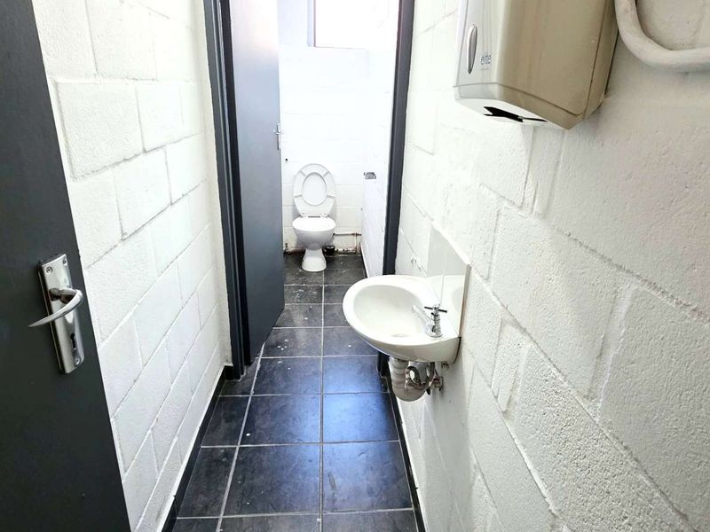 A functional industrial-style bathroom featuring a wall-mounted basin, toilet, tiled flooring, and exposed utility conduits.