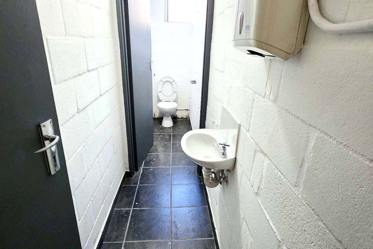 A functional industrial-style bathroom featuring a wall-mounted basin, toilet, tiled flooring, and exposed utility conduits.