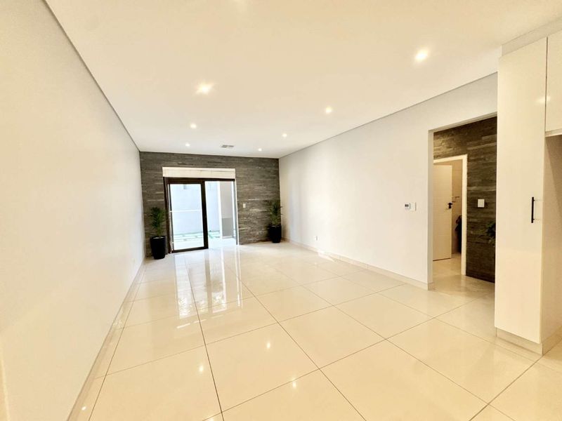 A bright, modern open-plan living area featuring polished porcelain tiles, recessed lighting, and a contemporary stone feature wall leading to an outdoor space.