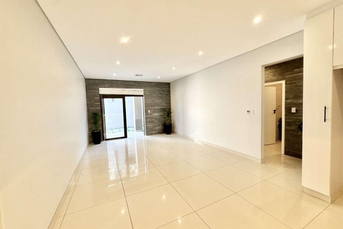 A bright, modern open-plan living area featuring polished porcelain tiles, recessed lighting, and a contemporary stone feature wall leading to an outdoor space.