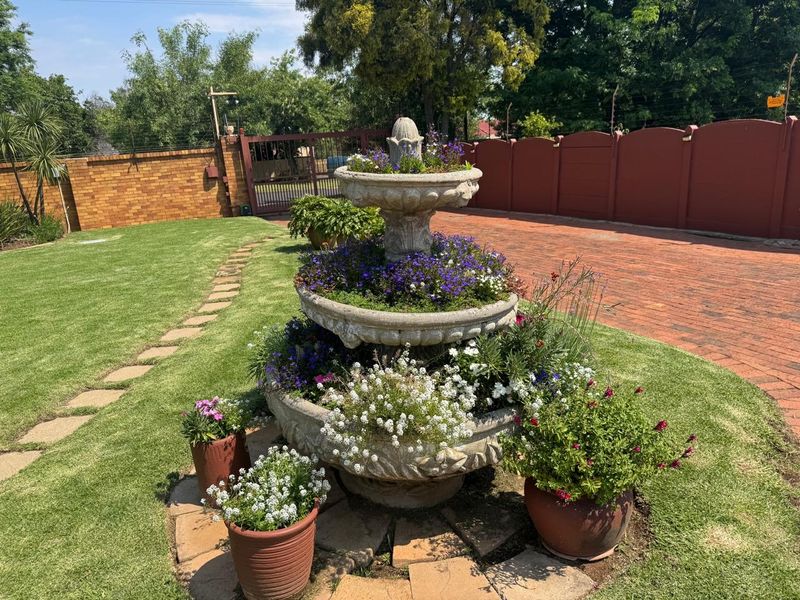 A well-maintained garden featuring a tiered stone fountain, lush lawn, and brick driveway enclosed by a secure perimeter wall.