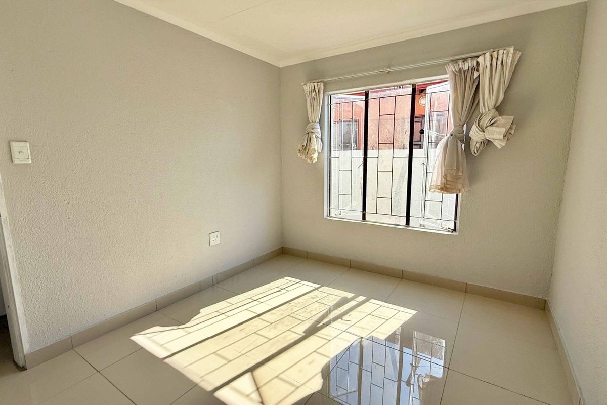 A bright, empty bedroom featuring neutral tiled flooring, a large window with security bars, and clean, minimalist wall finishes.