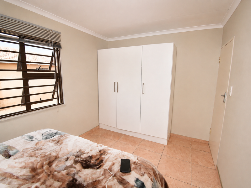 A bedroom featuring tiled flooring, a built-in wardrobe, and a window fitted with security bars and venetian blinds.
