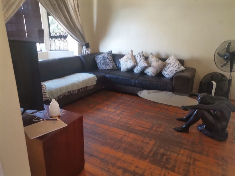 A living room featuring original hardwood flooring and windows fitted with security bars, offering a functional space with potential for cosmetic updates.