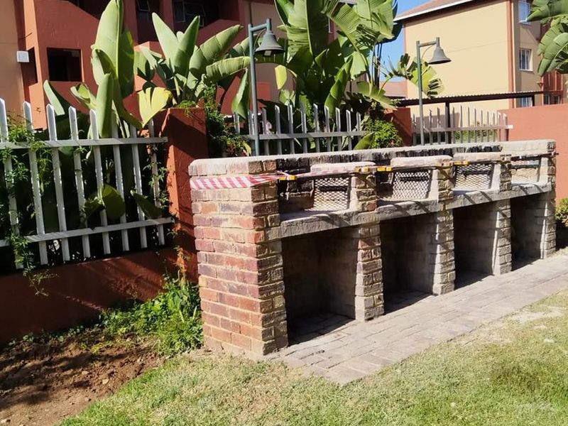 Communal outdoor area featuring a built-in brick braai station, paved walkway, and metal perimeter fencing set within a landscaped garden.